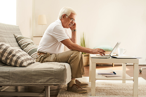 Side view portrait of modern senior man using laptop at home and speaking by phone