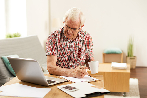 Portrait of senior man working with laptop at home counting finances and drinking coffee