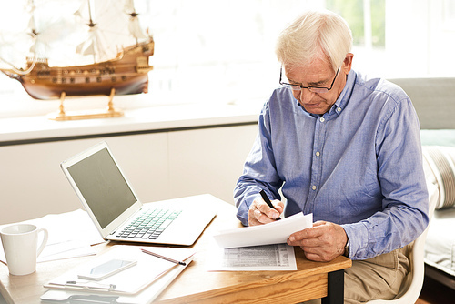 Portrait of modern senior man filling in application papers at home sitting at table with laptop in cozy living room