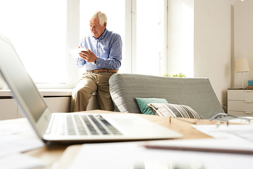 Portrait of modern senior sitting on windowsill at home, laptop in foreground