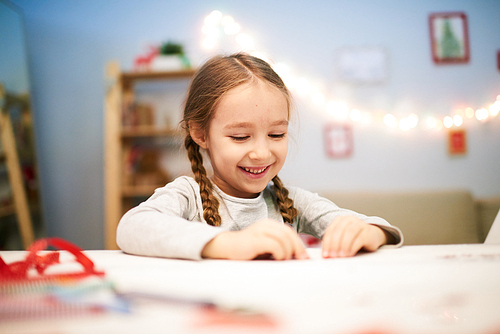 Cheerful little girl with toothy smile sitting at desk of cozy bedroom and preparing Christmas present for her mother, portrait shot