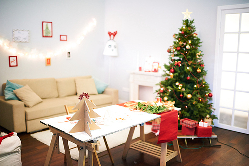 Preparations for Christmas celebration: cozy living room with festive Christmas tree, desk with unfinished decorations, handmade pictures hanging on wall