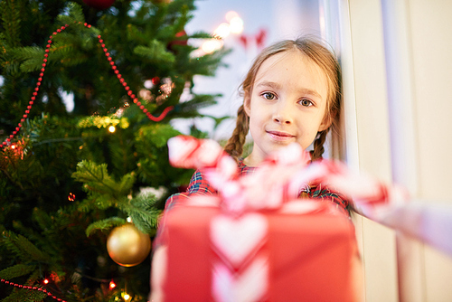 Portrait of cute little girl  with warm smile while passing Christmas present to someone, decorated Christmas tree on background