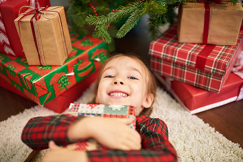Cute little girl posing for photography while lying on cozy carpet and embracing Christmas gift box, waist-up portrait