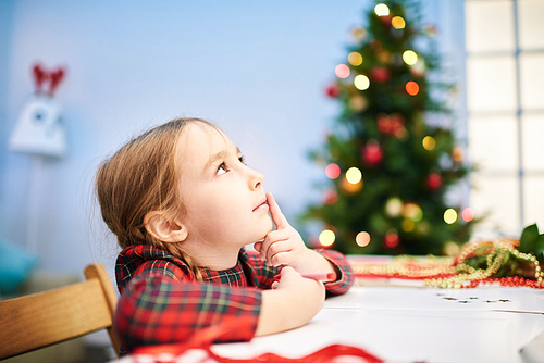 Profile view of pensive little girl looking away while thinking over design of future Christmas card for her grandmother, interior of cozy living room with Christmas tree on background