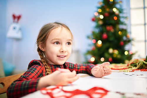 Creative little girl drawing Christmas card for her granny with wax crayons while sitting at desk of cozy living room, portrait shot