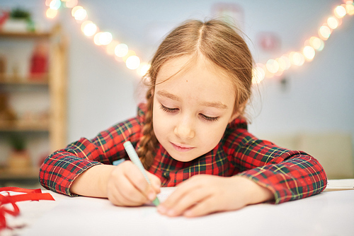 Head and shoulders portrait of little dark-haired girl wearing tartan dress wrapped up in drawing Christmas card while sitting at desk of cozy living room.