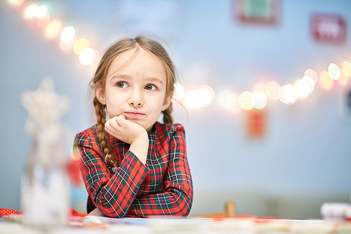 Waist-up portrait of thoughtful little girl wearing tartan dress resting head on hand while sitting at desk and thinking over Christmas present for her mom, blurred background