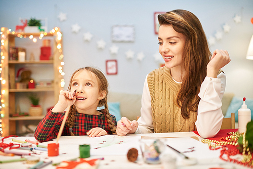 Dreamy little girl distracted from drawing colorful picture and looking upwards with interest, her attractive mother sitting next to her at desk, interior of decorated living room on background