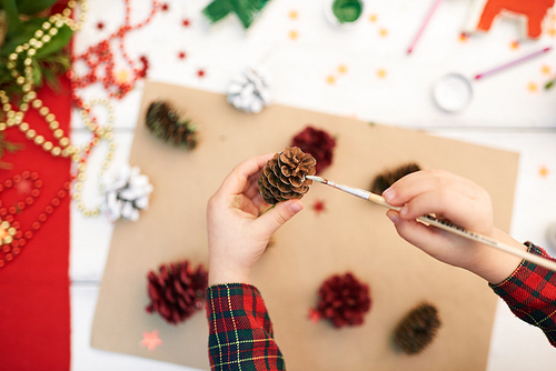 Close-up shot of unrecognizable little girl standing at wooden desk and covering pinecone with gouache while making decorations for Christmas