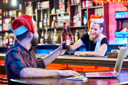 Portrait of bearded bartender wearing Santa hat cheering to customer with beer bottle in pub, celebrating Christmas, focus on foreground