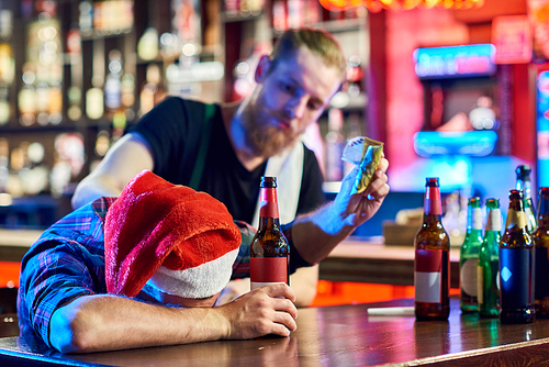 Portrait of drunk man wearing Santa hat laying on bar counter drinking beer and throwing money around after Christmas party in pub