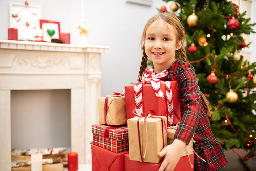 Portrait shot of adorable little girl wearing tartan dress  with toothy smile while gently embracing pile of Christmas gift boxes, interior of cozy living room on background