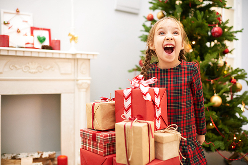 Portrait of cute little girl with two braids looking away and shouting with delight while  standing at pile of Christmas gift boxes, interior of decorated living room with fireplace on background