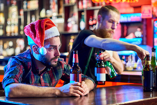 Portrait of young man wearing Santa hat getting drunk alone sitting at bar counter drinking beer at Christmas