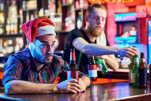 Portrait of young man wearing Santa hat getting drunk alone sitting at bar counter drinking beer at Christmas, copy space