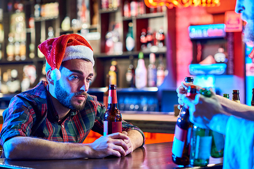 Portrait of sad young man wearing Santa hat getting drunk alone sitting at bar counter drinking beer at Christmas, copy space