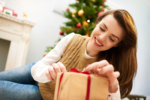 Cheerful woman with charming smile wearing knitted vest and jeans opening gift box while celebrating Christmas Eve at cozy living room, portrait shot