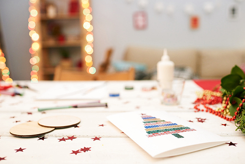 Close-up shot of wooden table surface: handmade Christmas card, pencils, wax crayons and confetti, blurred background