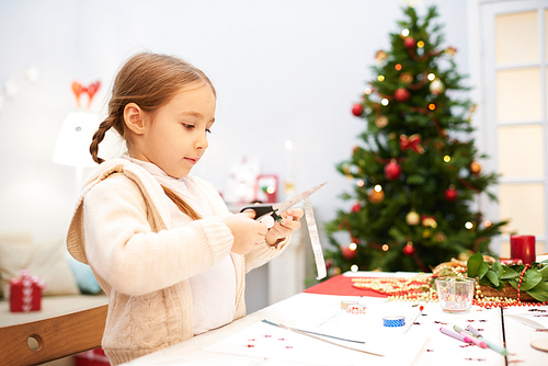 Cute little girl wearing knitted cardigan holding scissors in hand while wrapped up in making ideal Christmas card for dad, interior of cozy living room decorated for Christmas on background