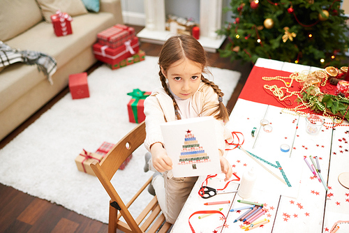 Portrait shot of pretty little girl showing handmade Christmas card to camera while standing at wooden desk covered with confetti, pencils and wax crayons