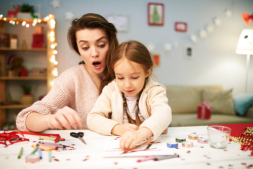 Surprised pretty woman keeping eye on her cute little daughter while making Christmas decorations together, interior of spacious living room on background