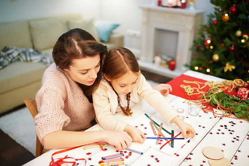 Lovely moments of motherhood: pretty woman wearing knitted sweater helping her little daughter to make Christmas decoration while gathered together at cozy living room