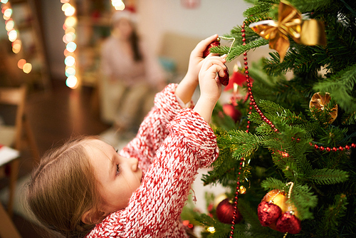 Portrait of dark-haired little girl wearing knitted sweater standing on toes while decorating Christmas tree, blurred background