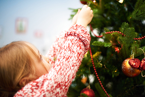 Close-up shot of adorable little girl wearing knitted sweater wrapped up in decorating Christmas tree while standing on toes, blurred background
