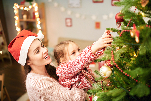 Profile view of attractive woman wearing knitted sweater and Santa hat holding her cute little daughter on arms while decorating Christmas tree together