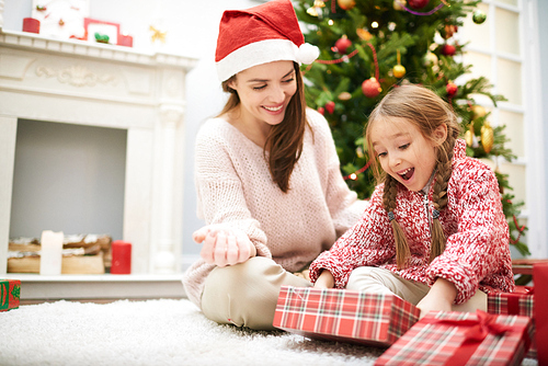 Cheerful little girl sitting by fireplace and unwrapping Christmas presents, her pretty mother wearing Santa hat looking at her with toothy smile