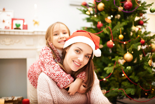 Family portrait of attractive woman wearing Santa hat sitting by fireplace while her cute little daughter embracing her tenderly, decorated Christmas tree behind  them