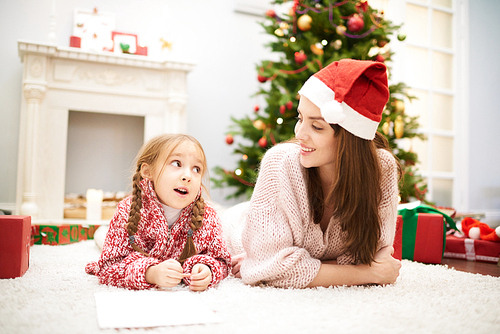 Lovely family portrait: adorable little girl wearing knitted sweater lying on cozy carpet by fireplace and chatting with her mom, interior of living room with decorated Christmas tree on background