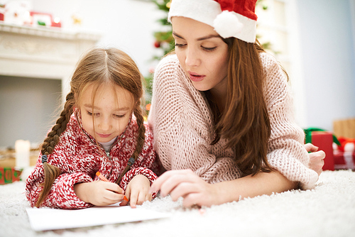 Making Christmas decorations together: cute little girl and her beautiful mother lying on cozy carpet and drawing colorful picture with help of wax crayons, interior of living room on background