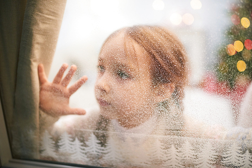 Pretty little girl wearing knitted sweater looking out window covered with frost while waiting for Santa Claus, portrait shot