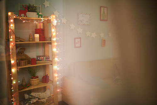 Interior of cozy living room decorated for Christmas celebration: small pictures hanging on wall, bookshelf with fairy lights, view through window