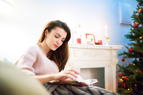 Portrait of attractive young woman wearing knitted sweater sitting on cozy sofa and reading adventure story, interior of cozy living room decorated for Christmas celebration on background