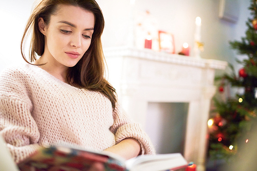 Waist-up portrait of attractive woman wearing sweater sitting on cozy sofa while wrapped up in reading interesting book, interior of living room decorated for Christmas celebration on background