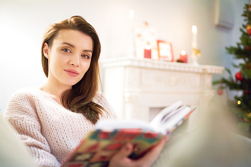 Beautiful young woman  with warm smile while distracted from reading adventure story, interior of cozy living room decorated for Christmas celebration on background