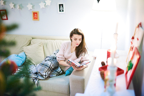 Pretty dark-haired woman sitting on cozy sofa and reading favorite book with interest while taking rest after celebrating Christmas with relatives