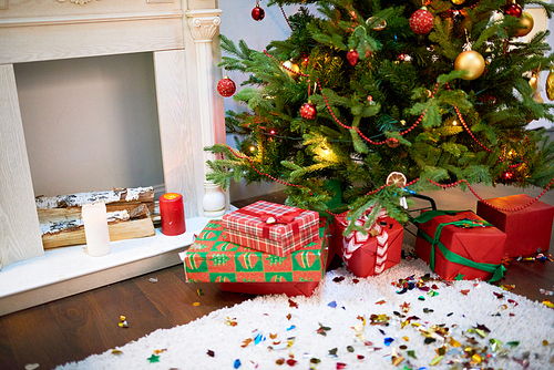 Interior of living room prepared for celebrating Christmas: pile of gift boxes lying under decorated Christmas tree, colorful confetti scattered everywhere, birch tree logs in fireplace