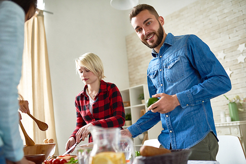Group of modern young people standing at big table with food on it preparing dinner together  at home, focus on smiling bearded man 