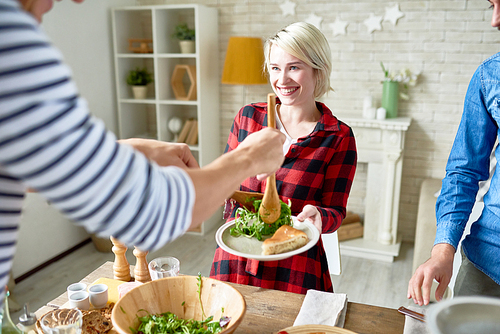 Group of modern young people standing at big table with food on it preparing dinner together, focus on happy blonde woman taking plate with pie