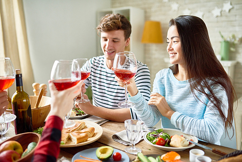 Group portrait of friends enjoying dinner together sitting at big table with delicious food , focus on young man and pretty Asian woman smiling happily clinking wine glasses celebrating holiday