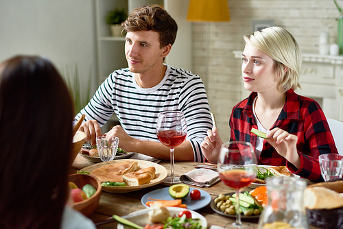Group portrait of  young friends having dinner together, gathered at big table with delicious dishes on it, focus on couple listening to conversation