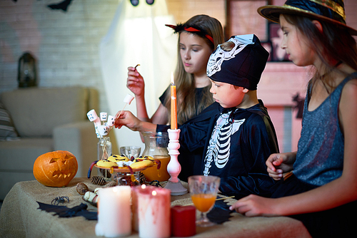 Portrait of three little kids wearing Halloween costumes standing at table with sweets during party in decorated room