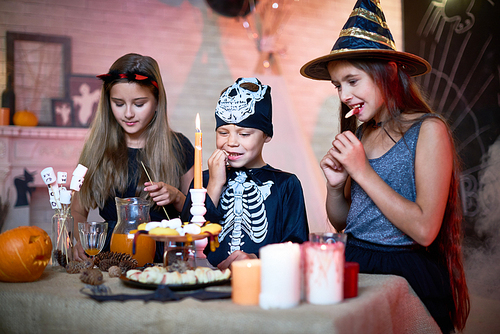 Portrait of three children wearing Halloween costumes eating sweets  and snacks during party in decorated room