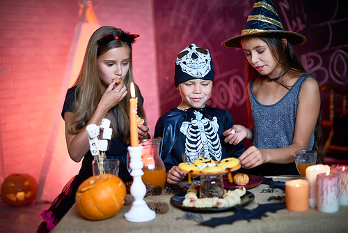 Positive curious kids eating sweet food from candy bar tasting Halloween treats in decorated studio at party