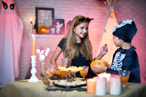 Portrait of two children, boy and girl wearing Halloween costumes chatting during party standing at table with sweets and snacks in decorated room