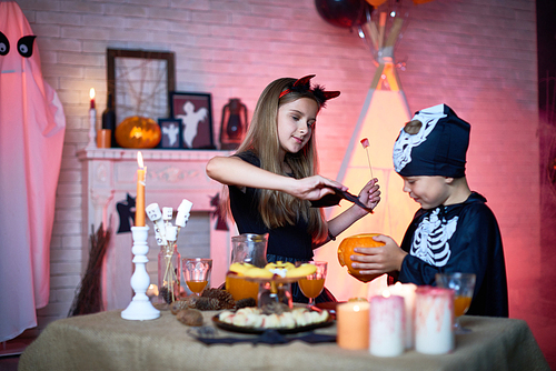 Portrait of children in Halloween costumes playing with carved pumpkin in decorated room during party